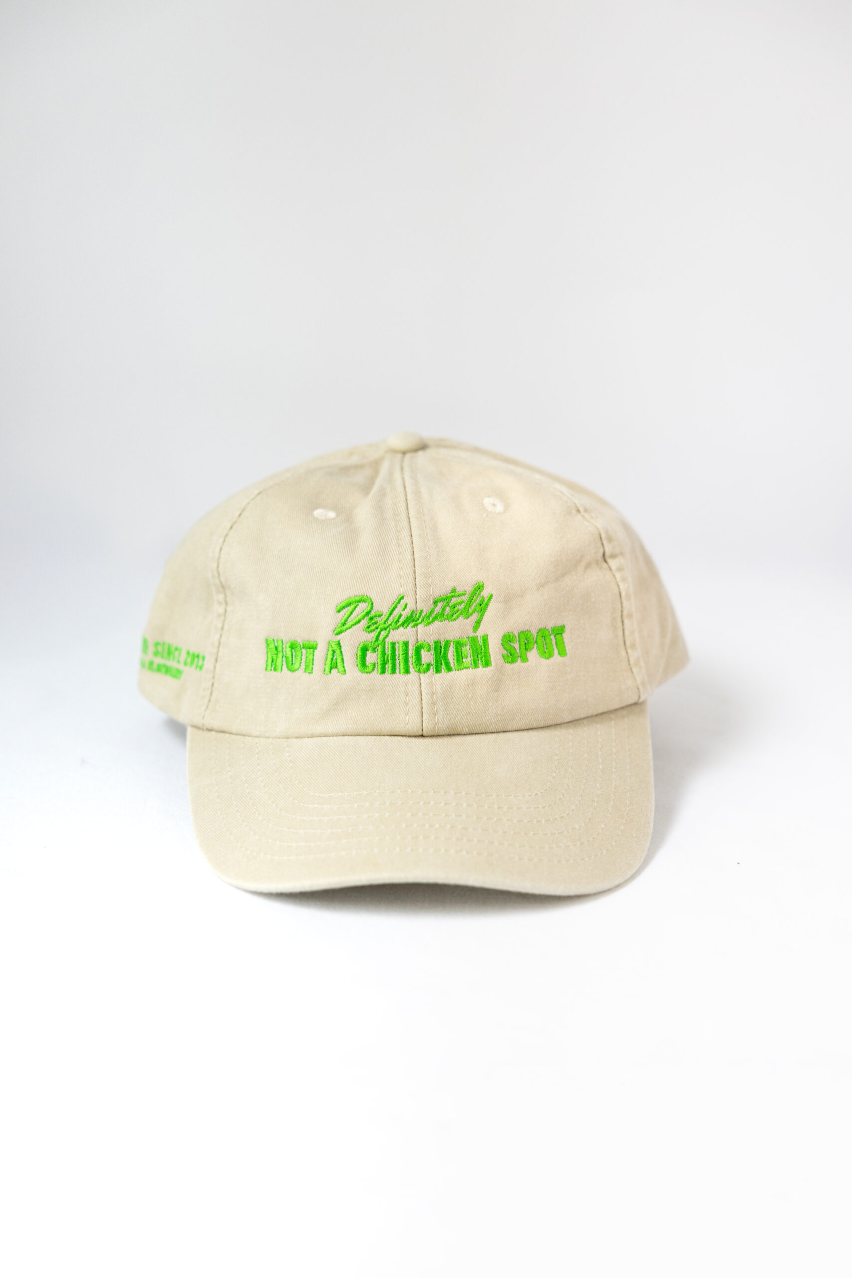 Le visuel montre une casquette beige de vue de face. Le style vestimentaire est décontracté et cool et l'on voit une broderie verte sur le devant indiquant « Definitely Not a Chicken Spot », un clin d'oeil au nom du traiteur Chicken Bacon Lettuce.