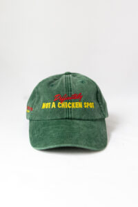 Le visuel montre une casquette verte de vue de face. Le style vestimentaire est décontracté et cool et l'on voit une broderie jaune et rouge sur le devant indiquant « Definitely Not a Chicken Spot », un clin d'oeil au nom du traiteur Chicken Bacon Lettuce.