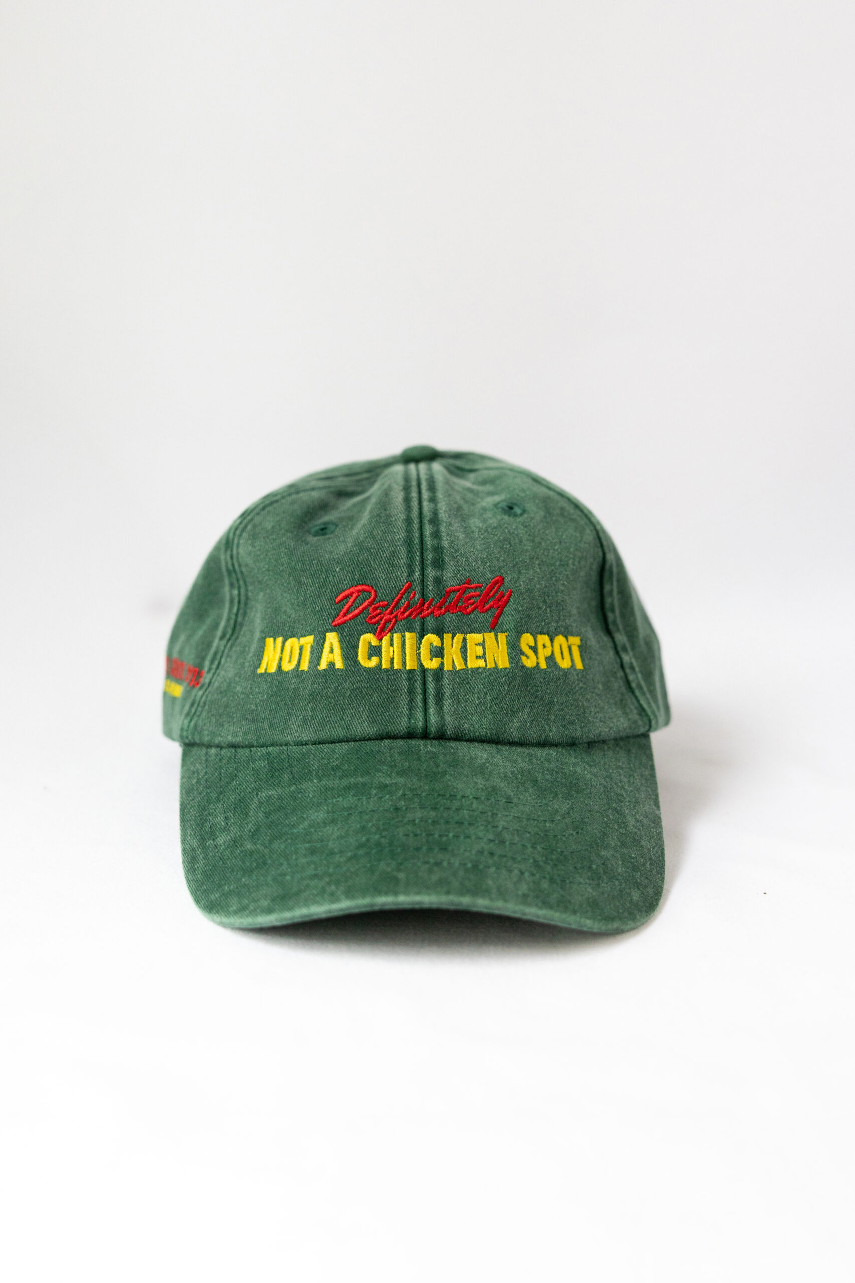 Le visuel montre une casquette verte de vue de face. Le style vestimentaire est décontracté et cool et l'on voit une broderie jaune et rouge sur le devant indiquant « Definitely Not a Chicken Spot », un clin d'oeil au nom du traiteur Chicken Bacon Lettuce.
