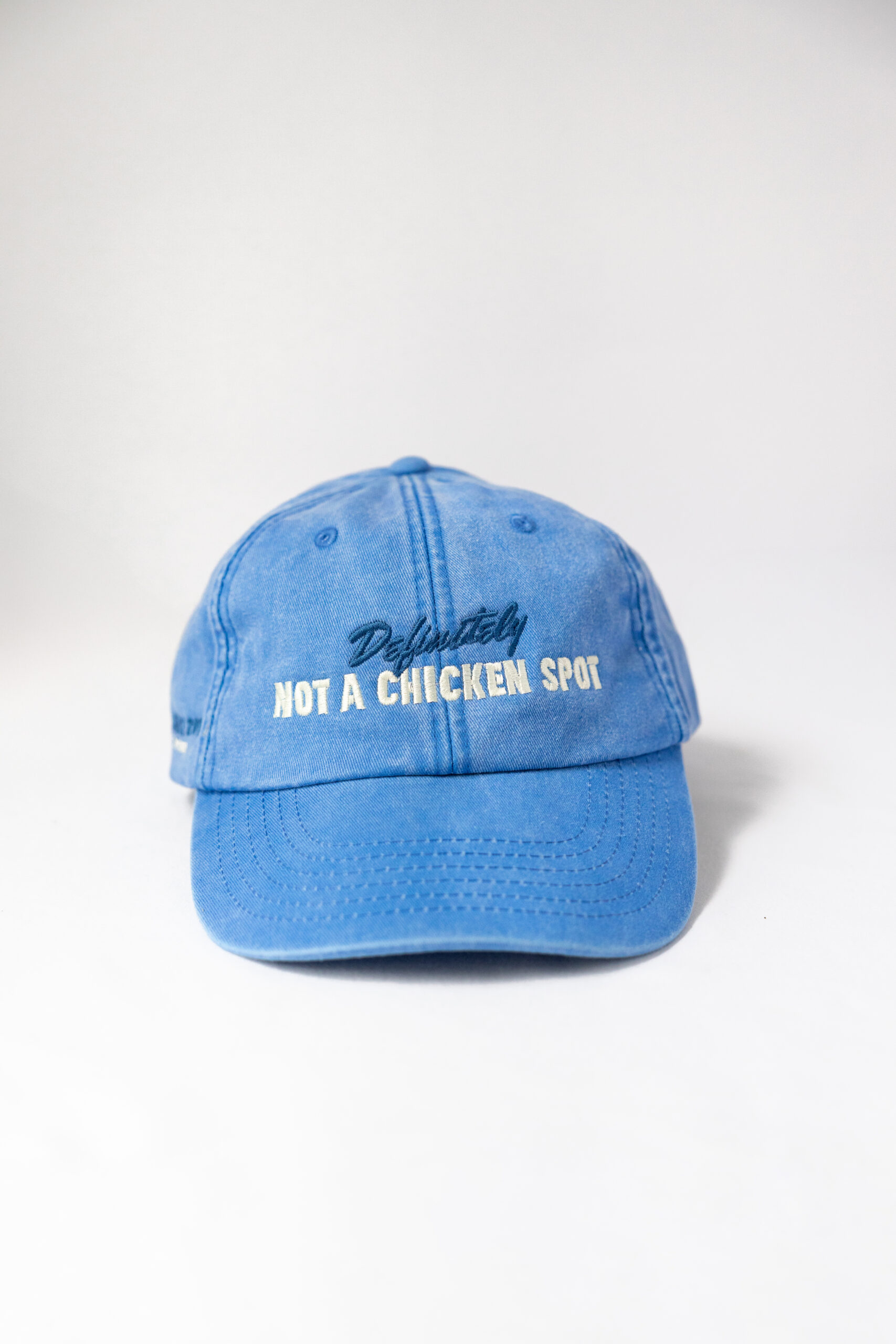 Le visuel montre une casquette bleue de vue de face. Le style vestimentaire est décontracté et cool et l'on voit une broderie blanc sur le devant indiquant « Definitely Not a Chicken Spot », un clin d'oeil au nom du traiteur Chicken Bacon Lettuce.
